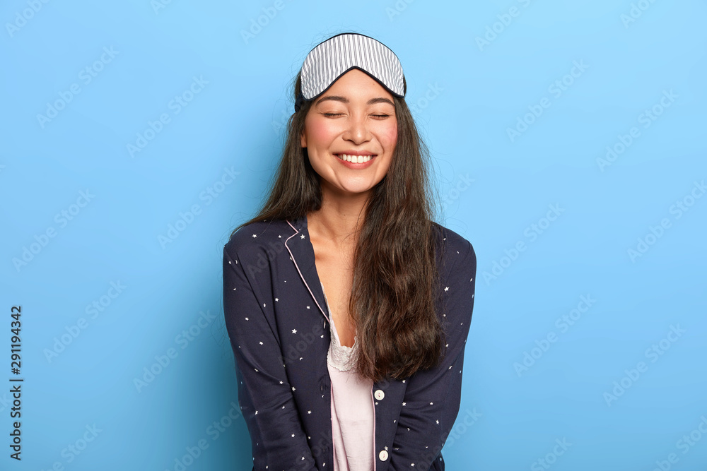 Indoor shot of cheerful woman with pleasant toothy smile, wears comfortable pyjamas and sleep mask, enjoys bedtime routine, smiles broadly at camera, poses in studio against blue studio wall Indoor shot of cheerful woman with pleasant toothy smile, wears comfortable pyjamas and sleep mask, enjoys bedtime routine, smiles broadly at camera, poses in studio against blue studio wall