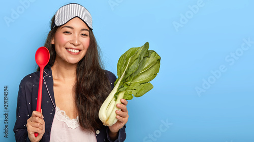 Photo of glad Asian woman has toothy smile, wears sleep mask and nightwear, poses with bok choy and red spoon, prepares tasty lunch isolated on blue wall copy space. Eating vegetables for good health