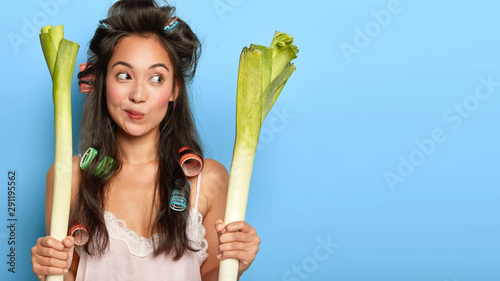 Thoughtful young female model going to make fresh salad and chop leek, purses lips, has long dark hair, dressed in nightwear, keeps to healthy diet, stands over blue wall. Organic food and eating