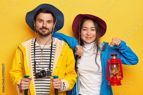 Horizontal shot of happy mixed race woman and man tourists spend summer vacation actively, walk on foot, cover long destination, male holds trekking sticks, retro camera, female with kerosene lamp photo