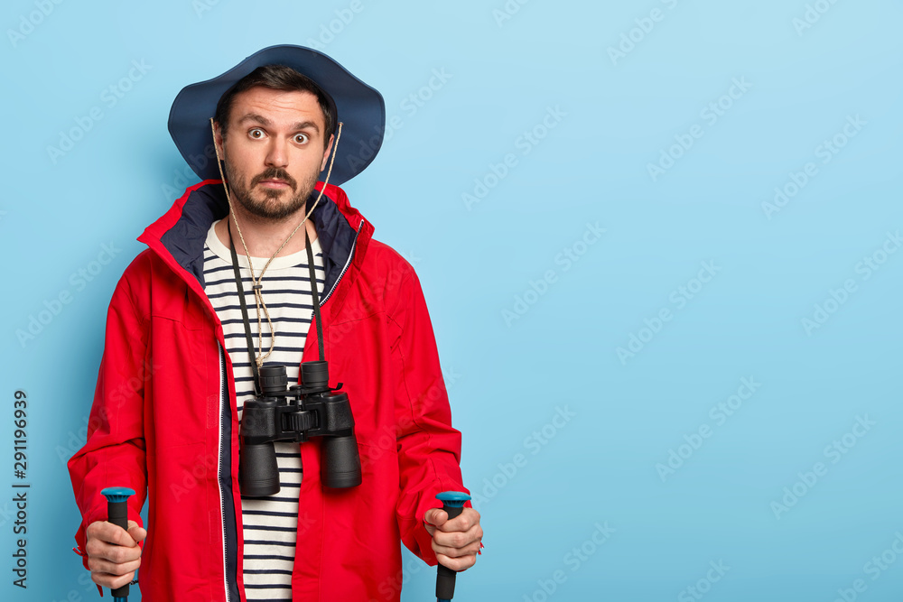 Camp and adventure concept. Surprised guy with mustache wears hat and red jacket, carries walking sticks, uses binoculars for exploration place, breathes fresh air, poses over blue wall being wondered Camp and adventure concept. Surprised guy with mustache wears hat and red jacket, carries walking sticks, uses binoculars for exploration place, breathes fresh air, poses over blue wall being wondered
