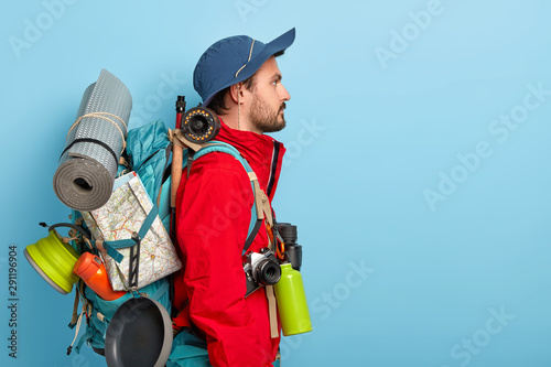 Profile shot of serious male backpacker stands with big rucksack, carries many necessary things for travelling and rest, goes camping alone, explores new surroundings, dressed in red jacket and hat