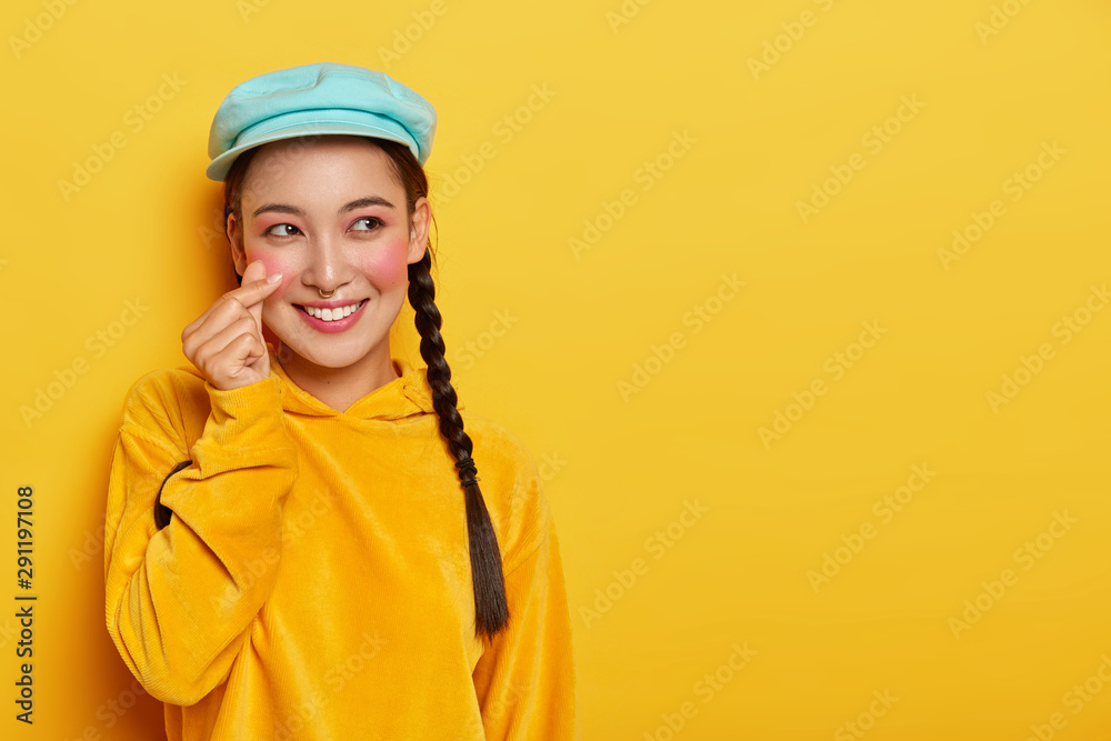 Smiling attractive Asain woman with pigtails, has rouge cheeks, makes korean like sign, wears beret and sweatshirt, has dreamy face expression, poses over yellow background, free space aside Smiling attractive Asain woman with pigtails, has rouge cheeks, makes korean like sign, wears beret and sweatshirt, has dreamy face expression, poses over yellow background, free space aside