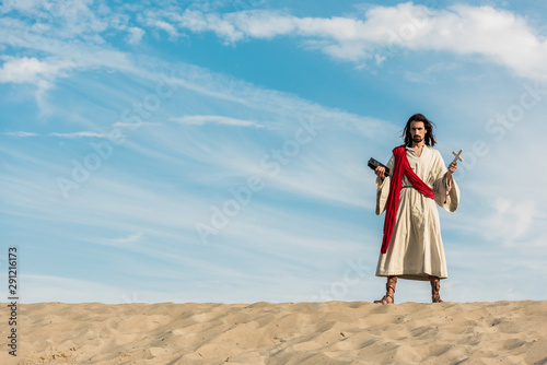 jesus holding holy bible and cross against blue sky with clouds in desert