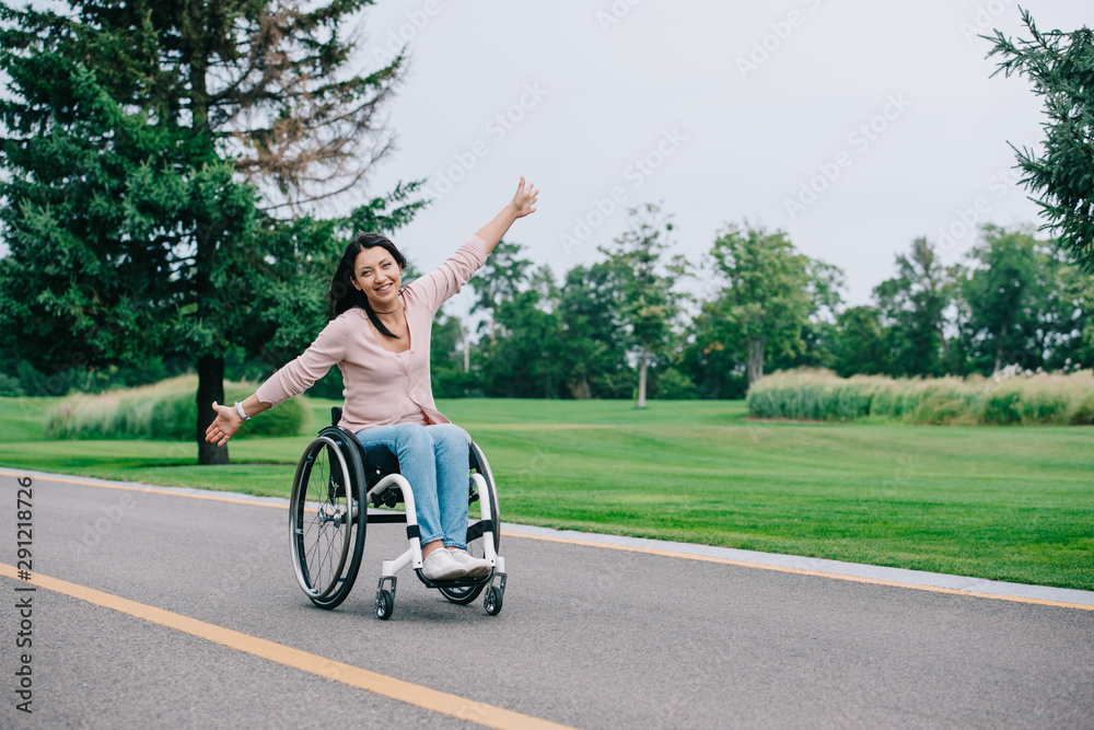 Fototapeta premium happy disabled woman gesturing and looking at camera while sitting in wheelchair in park