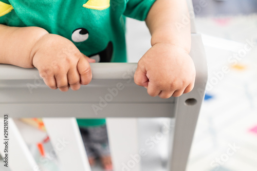 Close up of baby's hands while standing up and holding onto the railing of the crib. Wearing a cute green t-shirt in a bright minimalistic modern nursery bedroom.