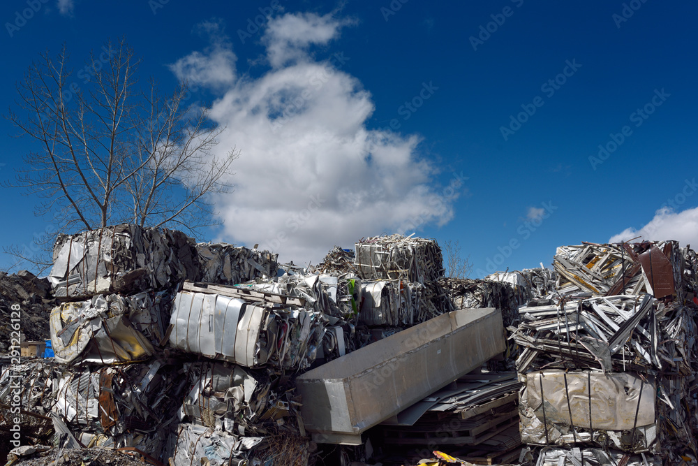 Piles of crushed cubes of recycled scrap metal with blue sky clouds and tree Piles of crushed cubes of recycled scrap metal with blue sky clouds and tree