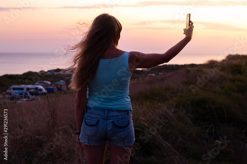 at sunset by the sea on a hill there is a girl in shorts and a T-shirt with a phone and takes a selfie
