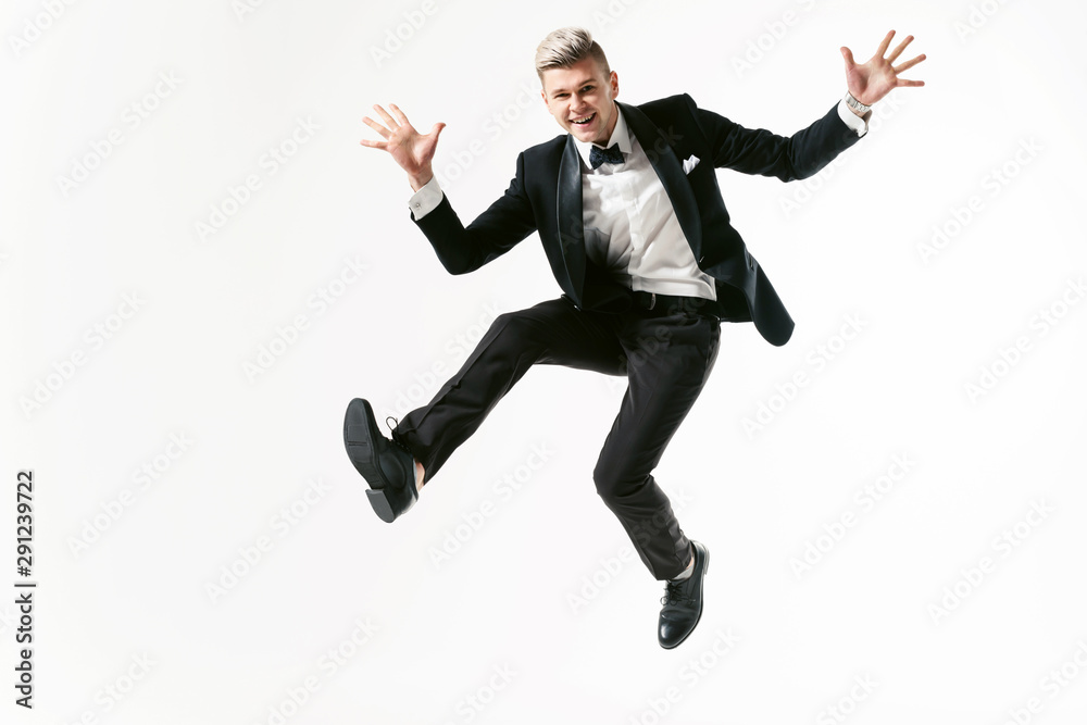 Portrait of young smiling handsome showman in tuxedo stylish black suit, studio shot jumping at white background. Businessman in jacket with bowtie