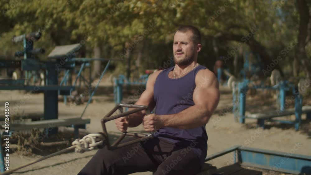 Concentrated confident fit man doing low pulley machine workout exercises in outdoor gym. Strong muscular bodybuilder practicing low cable pulley row seated while training on fresh air.