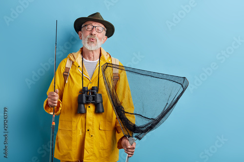 Tela funny old man with binocular, rod and net in yellow raincaot and grren hat blowing kiss, whistling close up photo