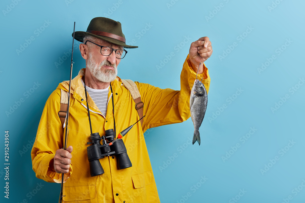 Stockfoto med beskrivningen Picture of unhappy handsome old fisherman with  blue eyes and beard holding fish, looking cranky and dissatisfied, unhappy  with poor catch. Hobby, leisure and occupation concept, fisherman