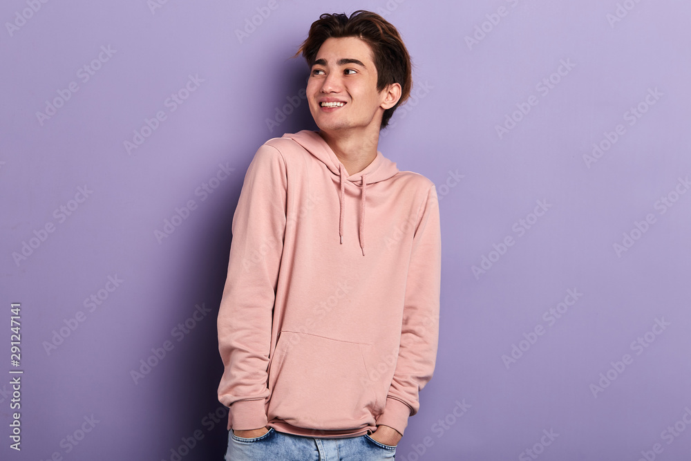 Relaxed young shy fashion man standing with hands on his pockets looking aside, careless life, isolated blue background, studio shot. . Relaxed young shy fashion man standing with hands on his pockets looking aside, careless life, isolated blue background, studio shot. .