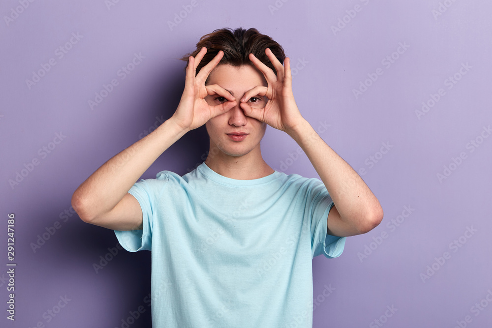 cheerful funny young man making binoculars with fingers wearing casual shirt isolated on blue background. close up portrait. cheerful funny young man making binoculars with fingers wearing casual shirt isolated on blue background. close up portrait.