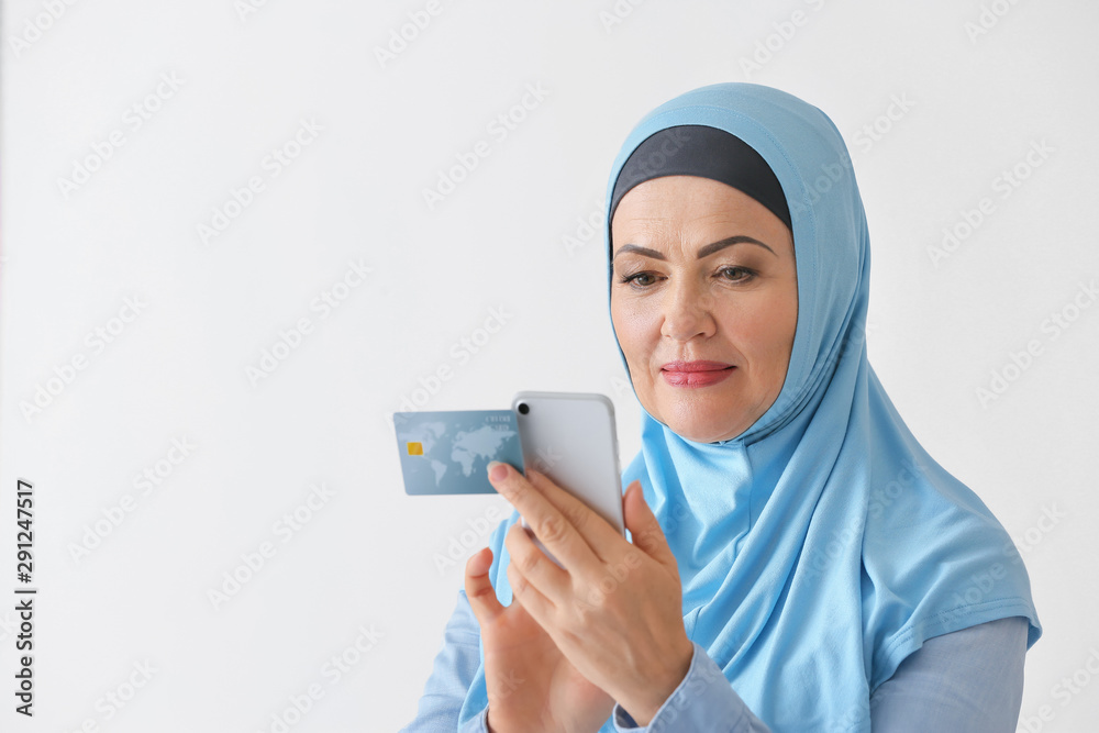 Beautiful mature Muslim woman with credit card and mobile phone on light background Beautiful mature Muslim woman with credit card and mobile phone on light background