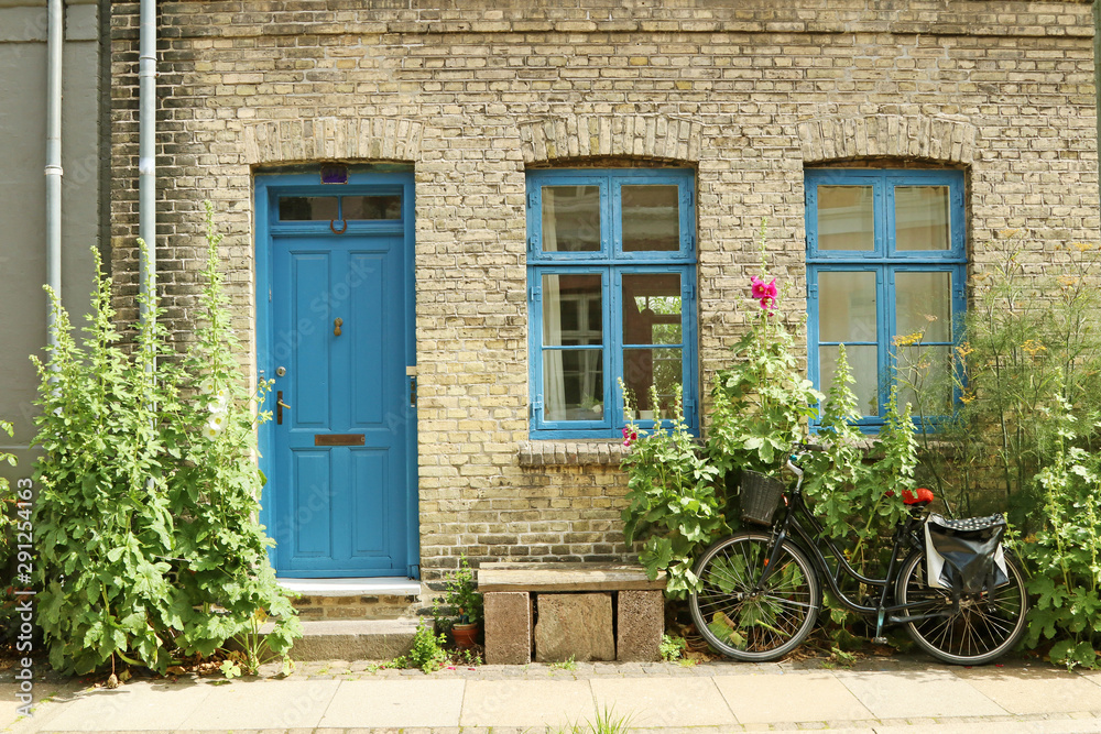 An old, gray house with a blue door and blue window frames. Hollyhock and dill grow near the wall.The bike is parked on the pavement. Bench and flower pot on the sidewalk.