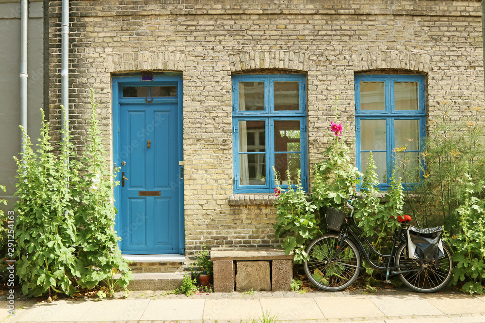 An old, gray house with a blue door and blue window frames. Hollyhock and dill grow near the wall.The bike is parked on the pavement. Bench and flower pot on the sidewalk. An old, gray house with a blue door and blue window frames. Hollyhock and dill grow near the wall.The bike is parked on the pavement. Bench and flower pot on the sidewalk.