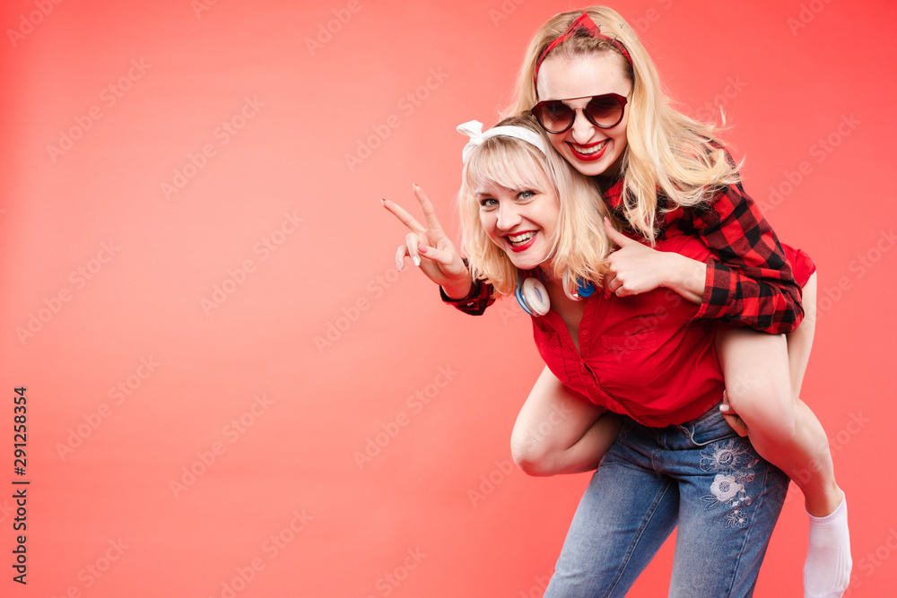 Stock studio portrait of two cheerful and positive trendy girlfriends or sisters having fun. Girl in sunglasses making bunny ears to her friend with her hand while sitting on her back. Isolate on red. - obrazy, fototapety, plakaty 