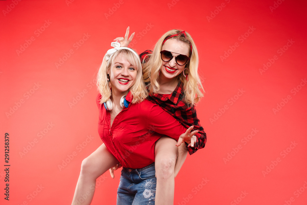 Stock studio portrait of two cheerful and positive trendy girlfriends or sisters having fun. Girl in sunglasses making bunny ears to her friend with her hand while sitting on her back. Isolate on red. - obrazy, fototapety, plakaty 