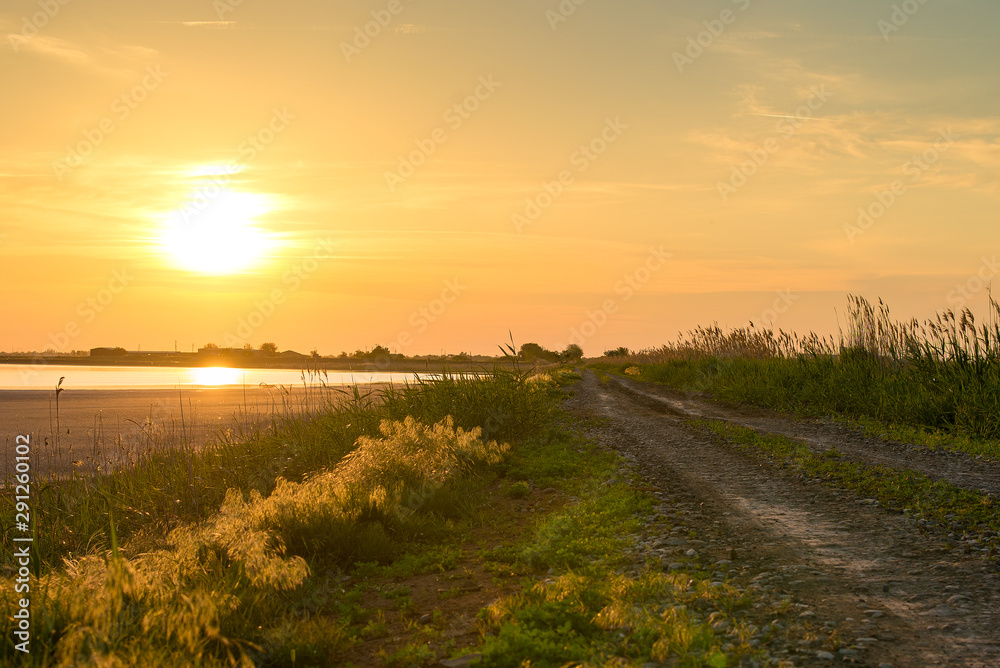 The road near the river goes into the distance. Sunset, road and lake.