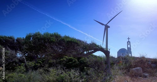 A modern windmill on the island of Crete near Elounda, against a background of blue sky and sea, with a small white church.