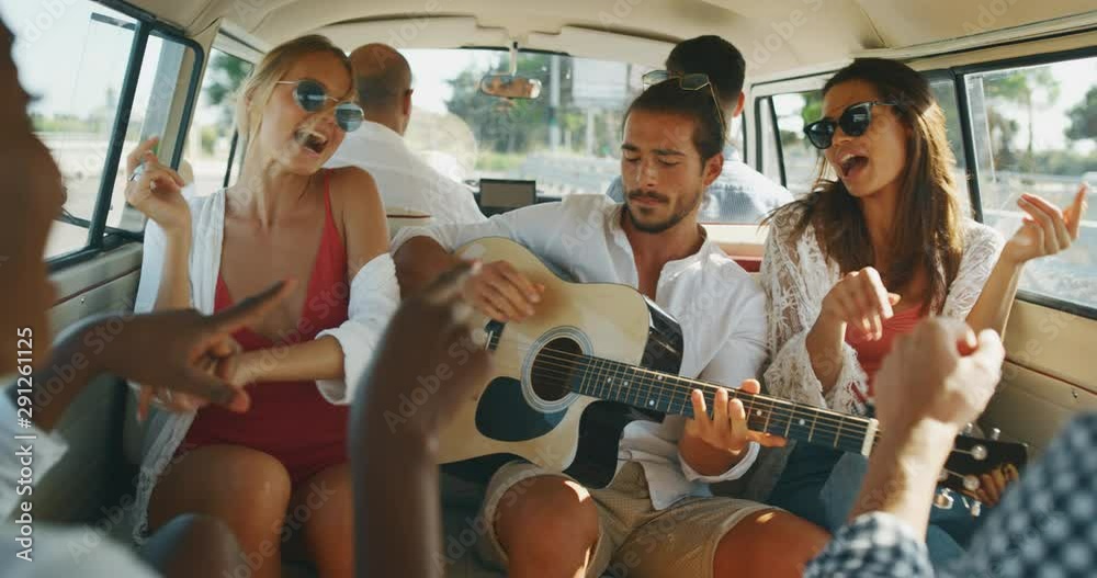 Slow motion of group of young multi-ethnic friends are having fun to play guitar, sing and dance in minivan during trip on coastal road along the sea.