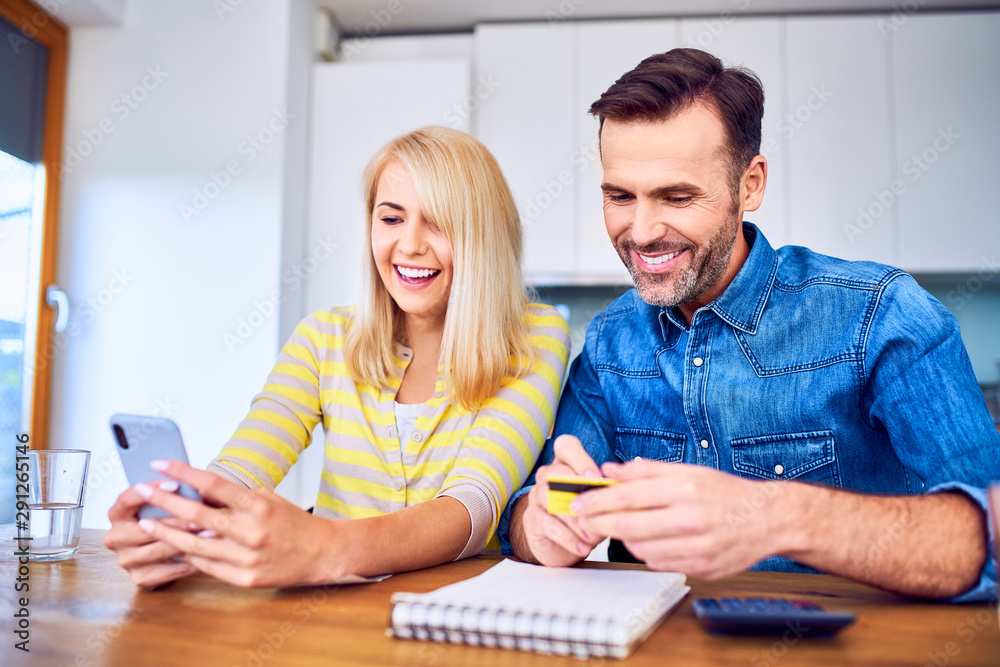 Joyful couple sitting at home paying online with smartphone and credit card Joyful couple sitting at home paying online with smartphone and credit card