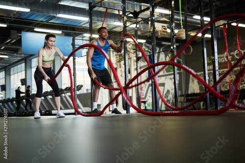 Wide angle portrait of fit young couple exercising with battle ropes during strength workout in cross functional gym, copy space