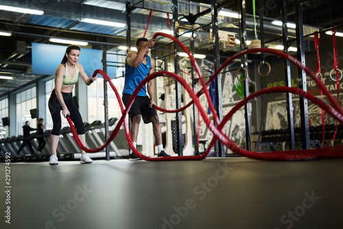 Wide angle portrait of fit couple exercising with battle ropes during strength workout in cross functional gym, copy space
