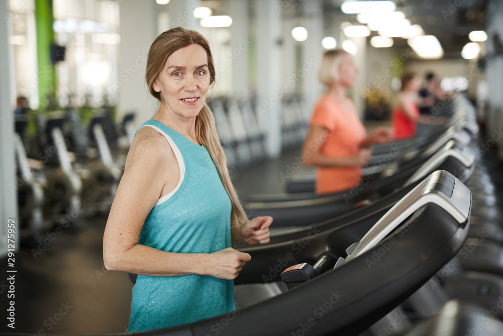 Waist up portrait of mature woman looking at camera while running on treadmill during cardio workout in modern gym, copy space