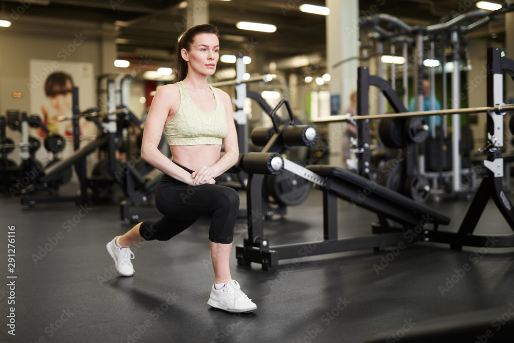 Full length portrait of fit young woman stretching legs during workout in modern gym, copy space Full length portrait of fit young woman stretching legs during workout in modern gym, copy space