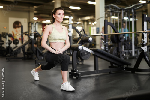Full length portrait of fit young woman stretching legs during workout in modern gym, copy space