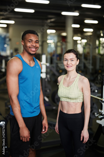 Portrait of fit young couple smiling at camera while posing standing in modern gym, fitness coach concept