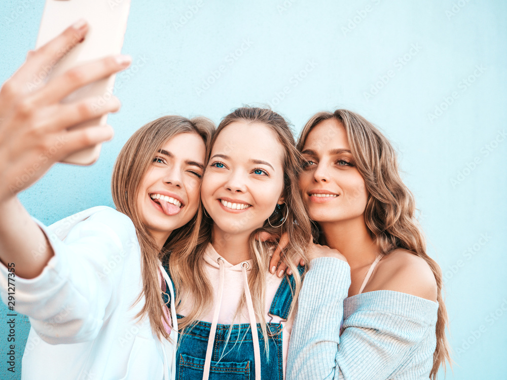 Three young smiling hipster women in summer clothes.Girls taking selfie self portrait photos on smartphone.Models posing in the street near wall.Female showing positive face emotions. Three young smiling hipster women in summer clothes.Girls taking selfie self portrait photos on smartphone.Models posing in the street near wall.Female showing positive face emotions.