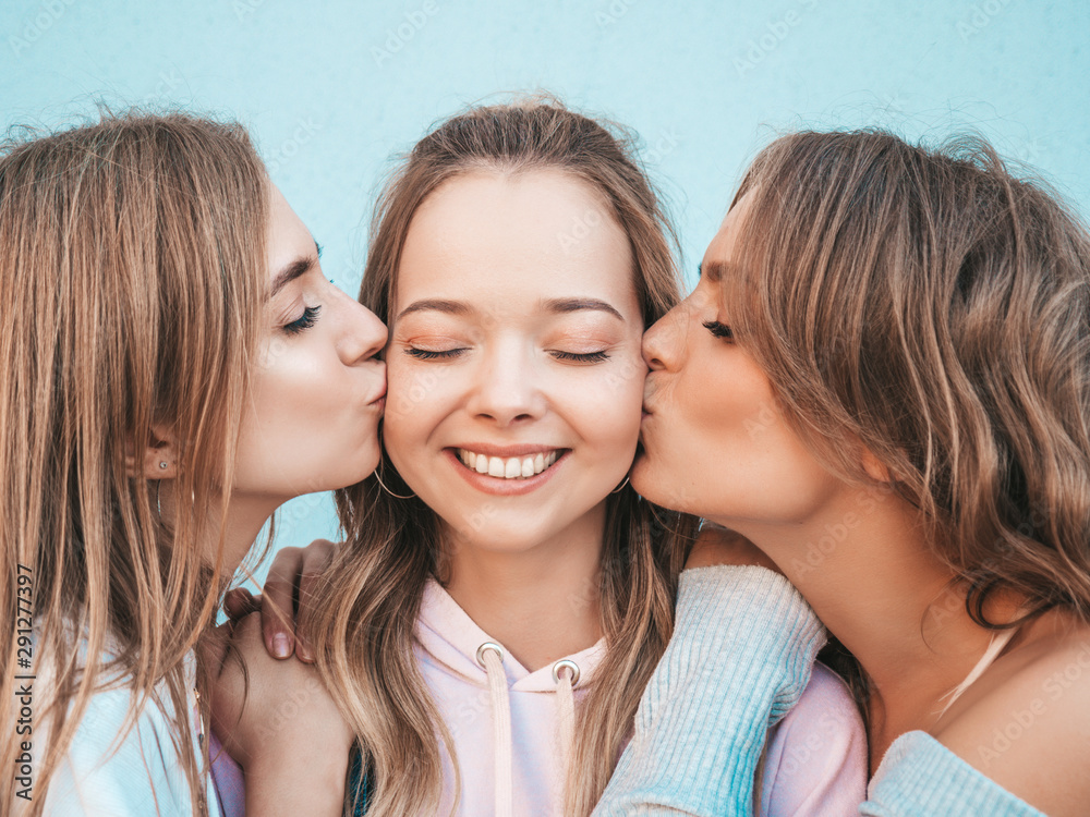 Portrait of three young beautiful smiling hipster girls in trendy summer clothes. Sexy carefree women posing in the street.Positive models kissing their friend in cheek