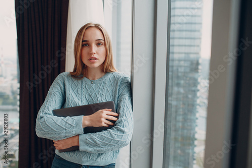 Free Download Portrait of stylish young woman in blue sweater with laptop standing by window - GFXLoad