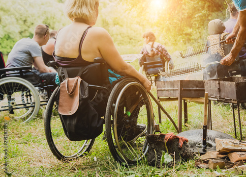 Disabled woman fries meat at the stake in a campsite with friends. Wheelchair in the forest on the background of bonfire. Barbeque. Camping.
