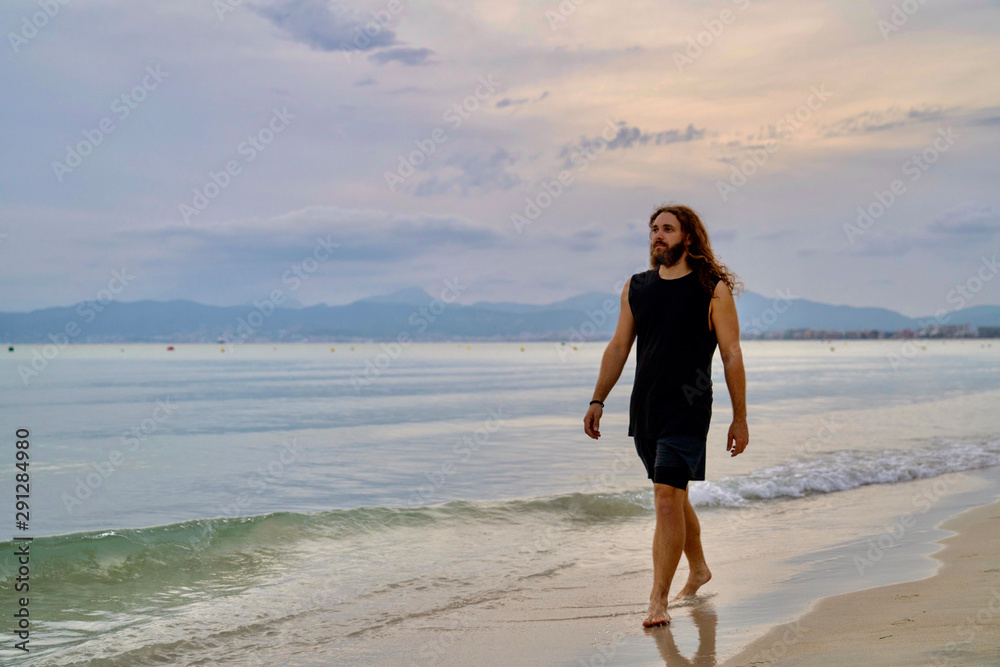 Model white caucasian bearded long haired man walks through sandy beach on sunset while a little wave breaks from right to left.