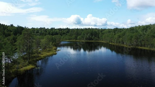 Flying over a beautiful small lake, with blue water surrounded by green forests. Summer landscape. Aerial view.