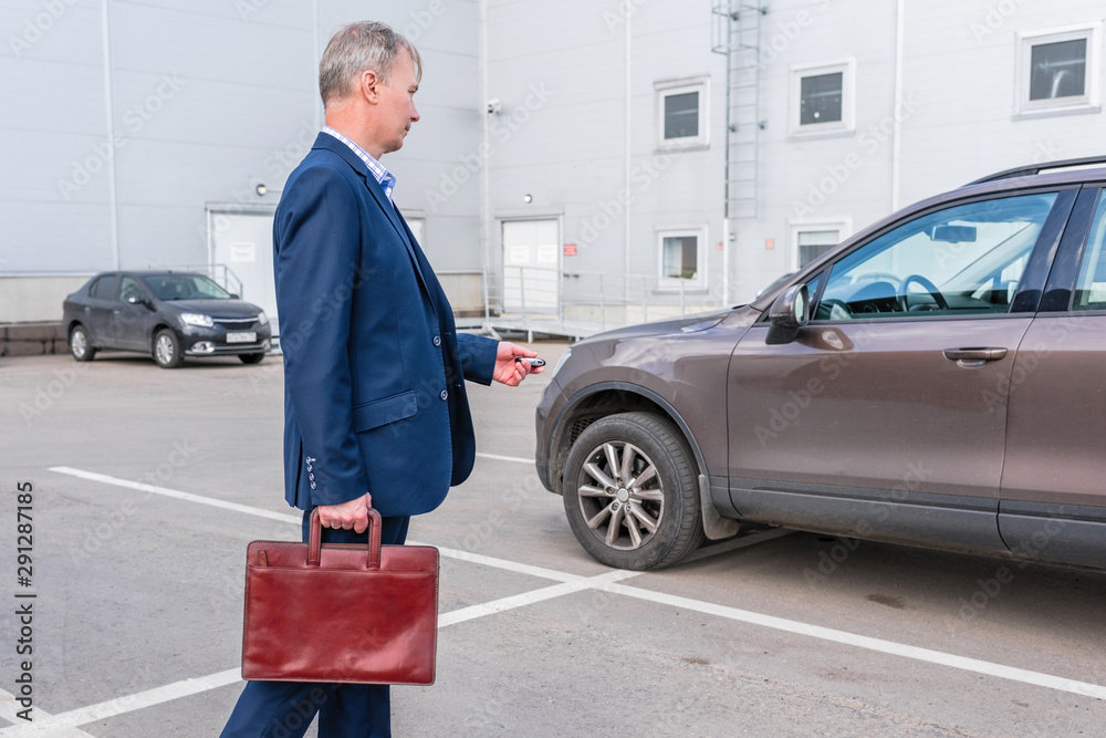 Gray-haired middle aged businessman in a blue suit and briefcase pushing button on remote control car key