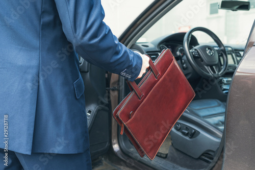 Middle-aged tall gray-haired businessman in blue suit with brown briefcase walks to his car on the private parking