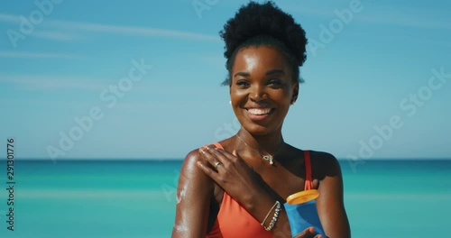 Slow motion of happy young african woman is applying a sunscreen or sun tanning lotion to take care of her skin during a vacation on a beach and smiling in camera.