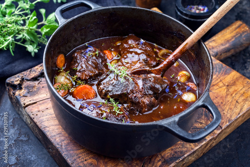 Traditionelle geschmorte deutsche Ochsenbäckchen in brauner Rotwein Sauce mit Karotten und Zwiebel angeboten als closeup in einem gusseisernen Schmortopf auf einem rustikalen Schneidebrett
