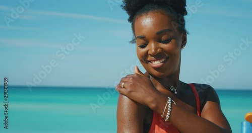 Slow motion of happy young african woman is applying a sunscreen or sun tanning lotion to take care of her skin during a vacation on a beach and smiling in camera. photo