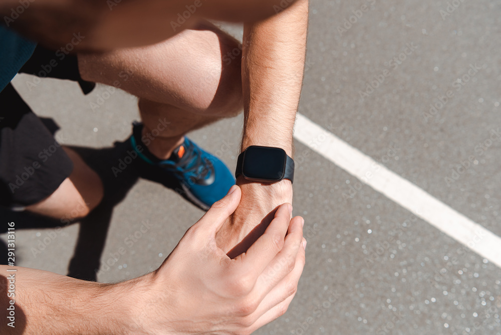 partial view of athletic young sportsman looking at smartwatch on running track at sunny day