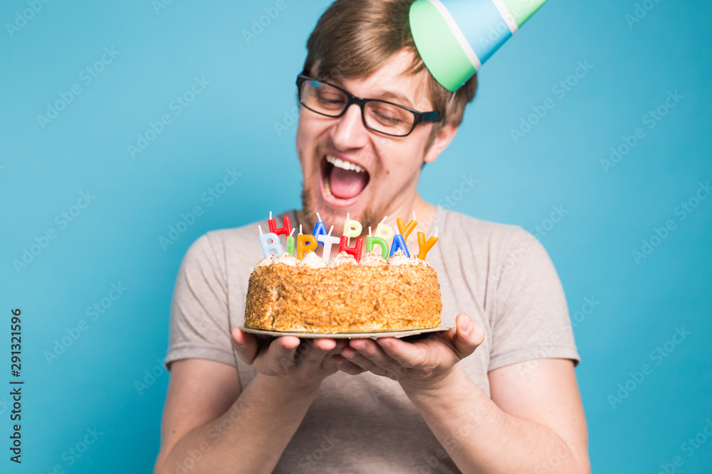 Funny male in paper congratulatory hat trying to bite off a cake with a happy birthday candles stand on a blue background. Funny male in paper congratulatory hat trying to bite off a cake with a happy birthday candles stand on a blue background.