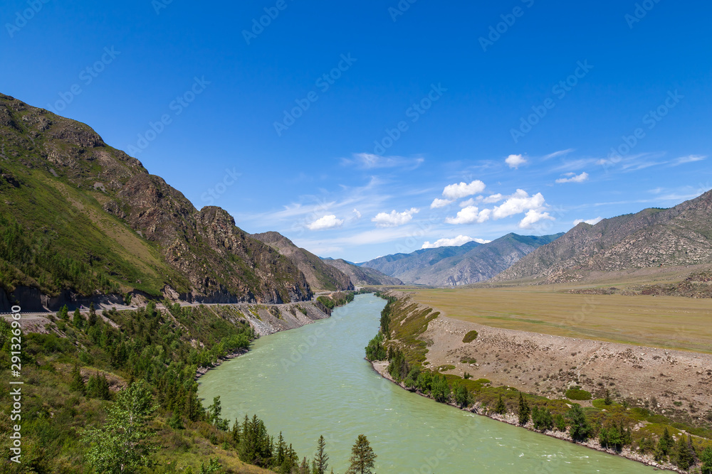 Landscape of the mountain chain of the Altai covered with green trees and rocks, with the turquoise Katun River and its rapids on a sunny summer day and a blue sky with white clouds. Tourist route.