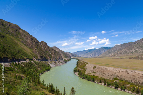 Landscape of the mountain chain of the Altai covered with green trees and rocks, with the turquoise Katun River and its rapids on a sunny summer day and a blue sky with white clouds. Tourist route.