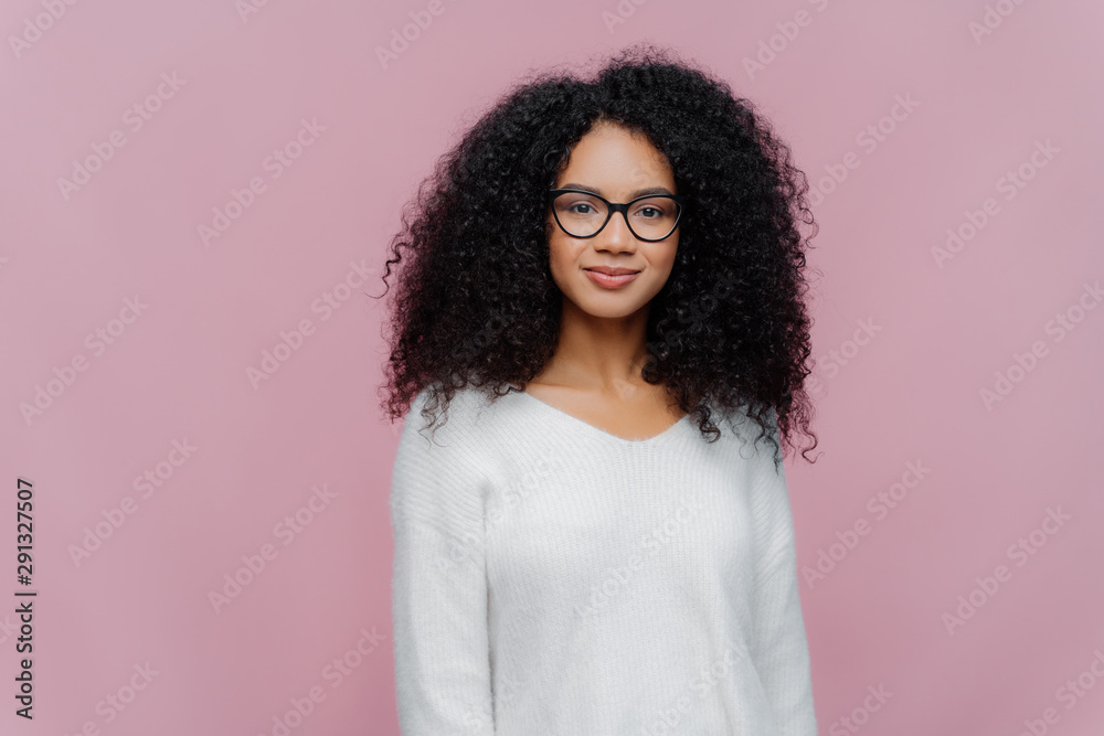 Half length shot of attractive African American woman looks through transparent glasses, white sweater, has serious calm expression, poses against violet studio wall. Facial expressions concept Half length shot of attractive African American woman looks through transparent glasses, white sweater, has serious calm expression, poses against violet studio wall. Facial expressions concept