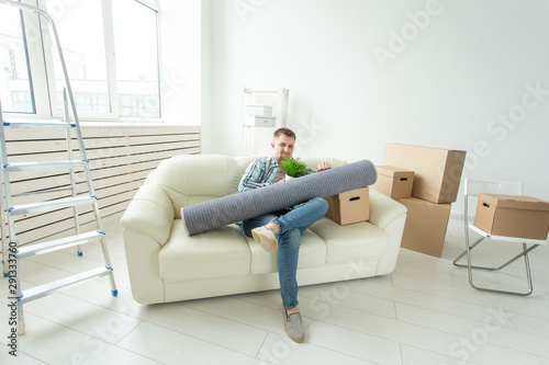 Positive young man sitting on the sofa with his things in the new living room after the move. Concept of housing affordability for young people. © satura_